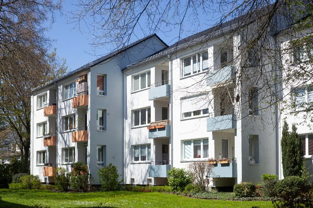 Gepflegtes Mehrfamilienhaus mit hellem Putz und Balkonen bei Sonnenschein (Symbolbild)