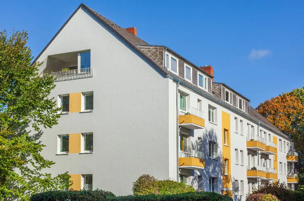 Weißes Mehrfamilienhaus mit gelben Fensterfeldern und Balkonen unter blauem Himmel, davor Herbstlaub — ein Haus, das ohne Inserat den Eigentümer wechseln kann